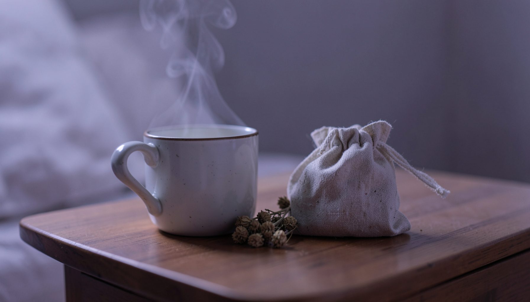 A steaming mug and a small linen pouch of dried valerian root on a bedside table, soft lavender-gray evening light